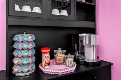 Cheerful pink coffee nook with a silver single-serve brewer, carousel of blue-labeled coffee pods, powdered creamer container and glass jars of sugar and sweeteners on a black countertop and shelving.