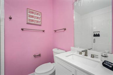 Bright pink bathroom interior with white vanity and sink, toilet, large mirror reflecting a glass-door subway-tile shower with built-in niche, brushed-nickel fixtures and playful “Howdy” wall art.