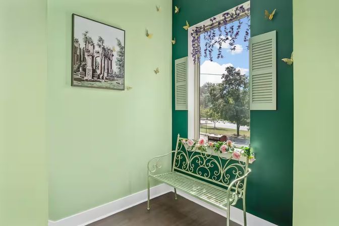 Cozy mint-green window nook with ornate metal bench decorated with pink roses beneath a shuttered window framed by hanging purple flowers, dark green accent wall with gold butterfly accents, framed classical artwork, and a tree-lined street view outside.