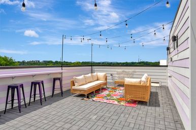 Sunlit rooftop patio with wicker sofas and beige cushions on a colorful floral rug, purple bar stools at a lavender railing, gray paver flooring, hanging string lights and a blue sky with distant treeline.