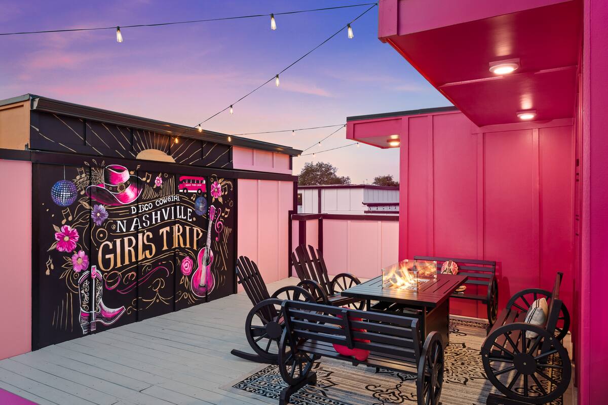 Dusk rooftop patio with hot-pink walls, string lights and wagon-wheel benches around a fire table, colorful mural reading Nashville Girls Trip with guitar and cowboy hat