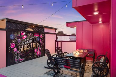 Dusk rooftop patio with hot-pink walls, string lights and wagon-wheel benches around a fire table, colorful mural reading Nashville Girls Trip with guitar and cowboy hat