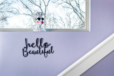 Lavender stairwell wall with a white bust wearing sunglasses and blowing a pink bubble, kiss mark on its cheek on the windowsill above a black script sign that reads "hello Beautiful," bare winter trees visible through the window.