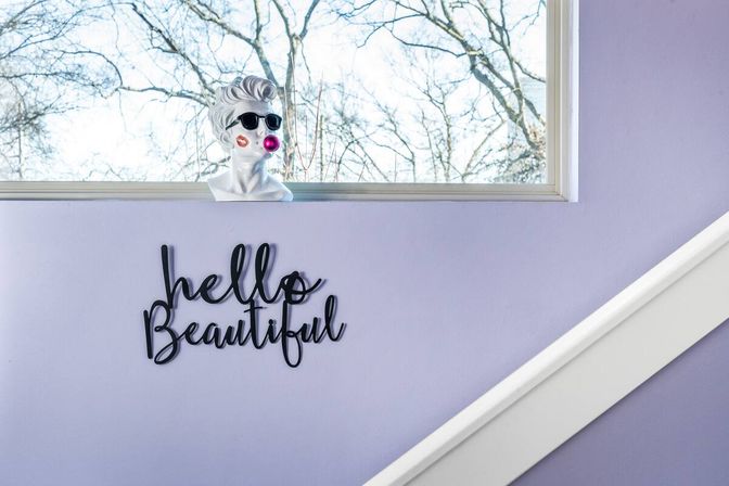 Lavender stairwell wall with a white bust wearing sunglasses and blowing a pink bubble, kiss mark on its cheek on the windowsill above a black script sign that reads "hello Beautiful," bare winter trees visible through the window.