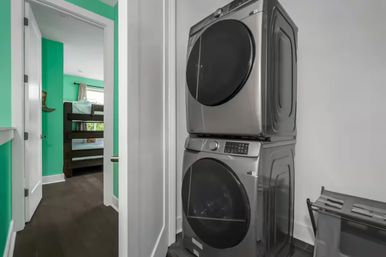 Stacked stainless steel front-load washer and dryer in a compact hallway laundry closet, with an open door revealing a bright teal-painted bedroom with a wooden bunk bed and dark hardwood floors.