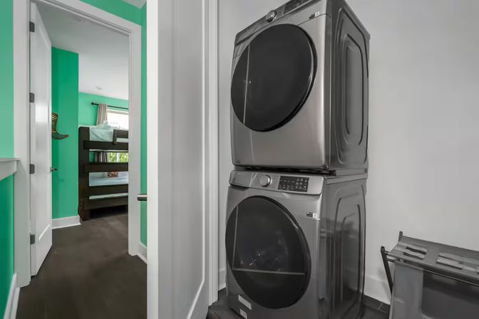 Stacked stainless steel front-load washer and dryer in a compact hallway laundry closet, with an open door revealing a bright teal-painted bedroom with a wooden bunk bed and dark hardwood floors.