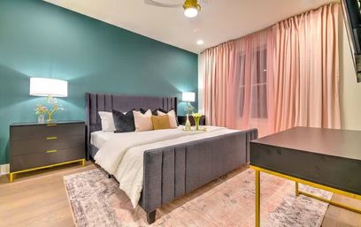 Cozy contemporary bedroom with a teal accent wall, gray upholstered bed dressed in white bedding and colorful throw pillows, pink floor-to-ceiling curtains, black nightstands and desk with gold accents, soft area rug and warm lamps.