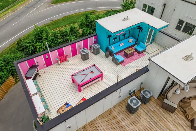 Aerial view of a vibrant urban rooftop terrace with a hot-pink pool table, teal lounge seating under a palm mural, pink-accented walls, outdoor bar stools, rug and fire table, and HVAC units overlooking a curved city road.