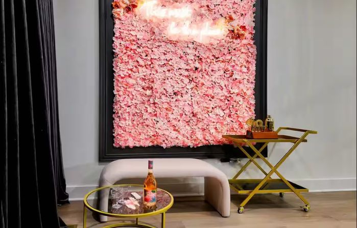 Stylish lounge interior with framed pink flower wall photo backdrop and glowing neon sign, blush curved bench, round glass-top coffee table with a bottle and glasses, gold rolling bar cart, black velvet curtain and light wood floor.