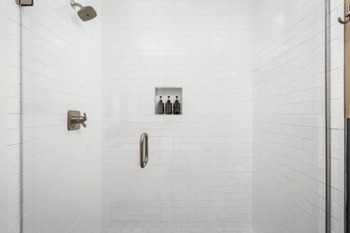 Crisp white subway-tile walk-in shower with glass door, brushed-nickel showerhead and handle, recessed niche holding dark shampoo bottles — modern bathroom