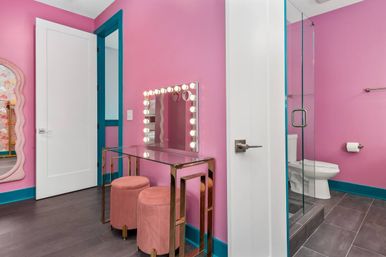 Playful bright pink makeup vanity and bathroom with teal trim, Hollywood mirror lights, glass shower, pink velvet stools and gray tile floor.