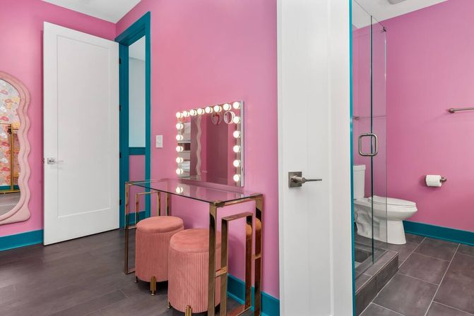 Playful bright pink makeup vanity and bathroom with teal trim, Hollywood mirror lights, glass shower, pink velvet stools and gray tile floor.