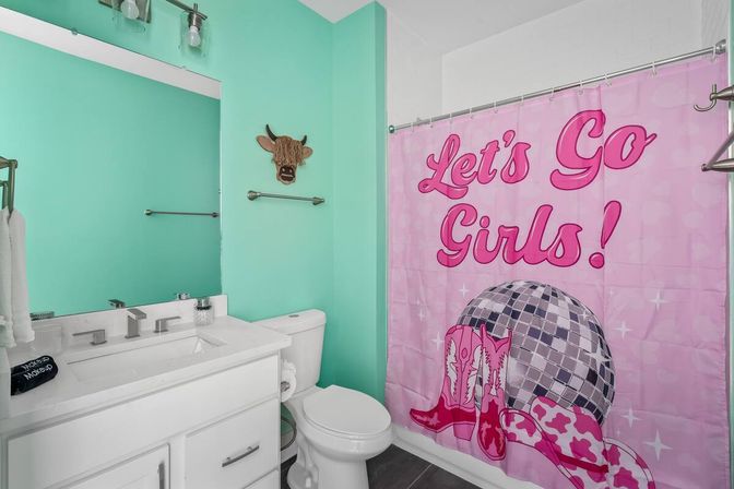 Bright turquoise bathroom with white vanity and toilet, cow wall decor and pink "Let's Go Girls!" shower curtain featuring a disco ball and cowboy boots.