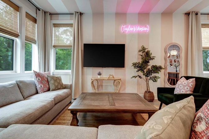 Cozy modern living room with beige striped accent wall and pink neon sign above a wall-mounted TV, large windows with bamboo shades, neutral sectional sofas, wooden coffee table, green velvet armchair, and potted plant.