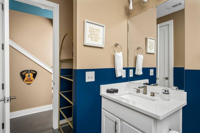 Modern residential bathroom with white vanity and quartz sink, large mirror and brushed-nickel faucet, beige upper walls with navy-blue wainscoting, twin towel rings with white towels, built-in open shelving, and inspirational wall art visible through an open doorway.
