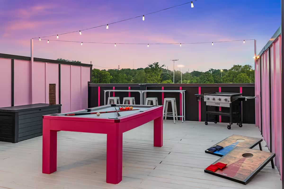 Rooftop patio game area at sunset with a bright pink pool table, cornhole boards, metal bar stools, grill, and string lights above overlooking treetops.