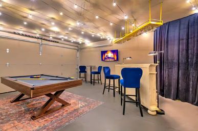 Cozy garage-style game room with a wooden pool table on a patterned rug, blue bar stools at a small lighted bar, wall-mounted TV showing a fireplace, hanging glass rack, and string lights across the ceiling.