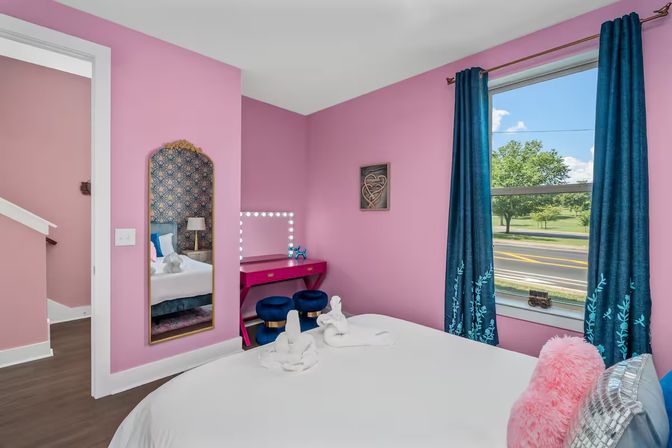 Playful bright pink bedroom with a white bed topped by towel swans, hot-pink vanity with illuminated mirror and blue stools, gold-framed floor mirror reflecting a patterned headboard, and teal curtains framing a window overlooking a tree-lined street.