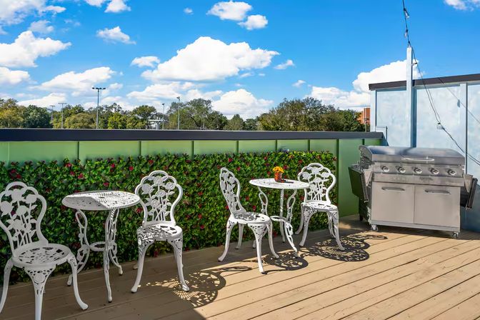 Sunny rooftop patio deck with white wrought-iron bistro tables and chairs, stainless-steel gas grill, faux green hedge and bright blue sky with puffy clouds.