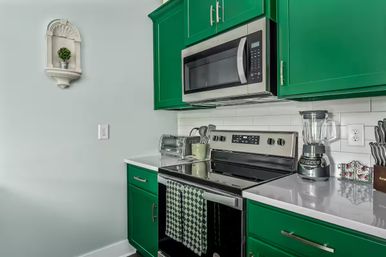 Modern kitchen with vibrant green cabinets, white subway tile backsplash, stainless steel microwave and electric stove, quartz countertops topped by a blender and toaster, black-and-white checkered dish towel and small wall shelf plant.