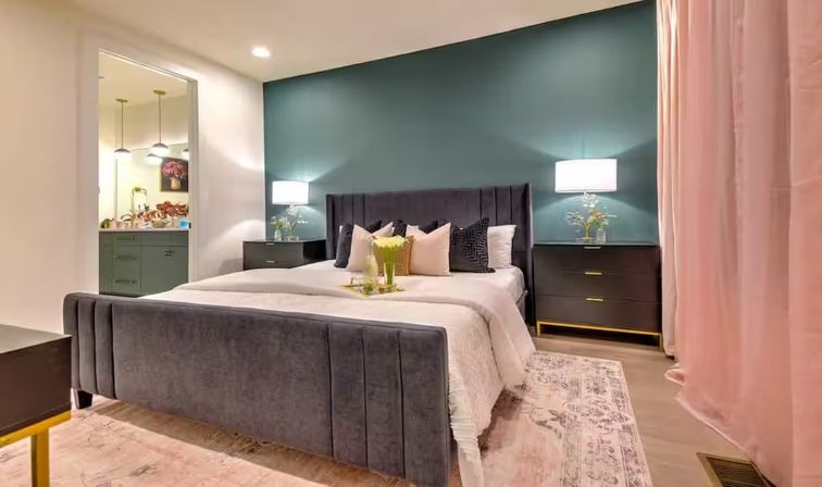 Stylish modern bedroom interior with a teal accent wall, gray velvet upholstered bed, matching nightstands with lamps, pink curtains, patterned area rug, and a visible en-suite vanity.