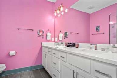Cheerful pink bathroom with white double-sink vanity, large mirror and exposed-bulb vanity lights, gray tile floor, glass shower and teal baseboard.