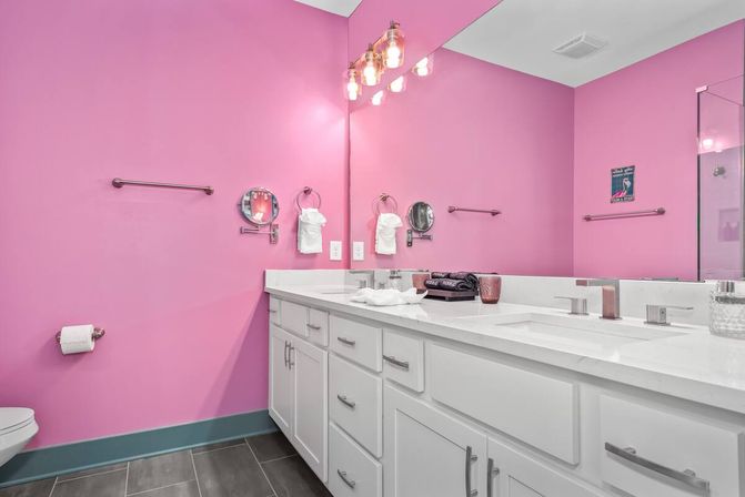 Cheerful pink bathroom with white double-sink vanity, large mirror and exposed-bulb vanity lights, gray tile floor, glass shower and teal baseboard.