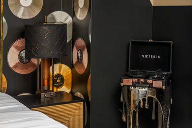 Black-and-gold bedroom corner with a vintage suitcase turntable on a dripping-paint stand, gold vinyl record wallpaper and a geometric black lamp on a nightstand.