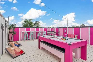 Playful rooftop patio game deck with a bright pink pool table, cornhole boards, mint-green bar stools, grill, string lights and palm decor under a sunny blue sky.