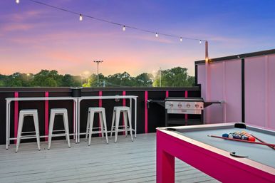 Pink-accented rooftop deck at sunset with string lights, high bar stools and counter, stainless-steel grill, and a bright pink pool table with racked billiard balls overlooking treetops.