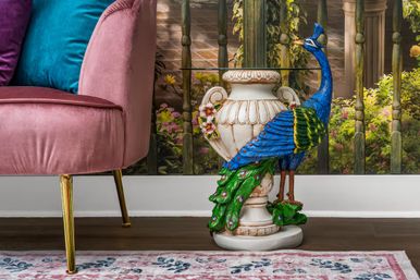 Colorful peacock sculpture perched on an ornate ceramic urn used as a side table beside a pink velvet sofa with a teal pillow, set on hardwood floors and a floral rug in a cozy living room.