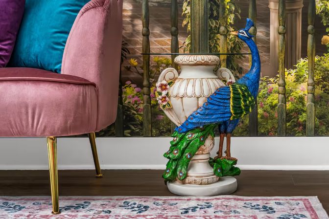 Colorful peacock sculpture perched on an ornate ceramic urn used as a side table beside a pink velvet sofa with a teal pillow, set on hardwood floors and a floral rug in a cozy living room.