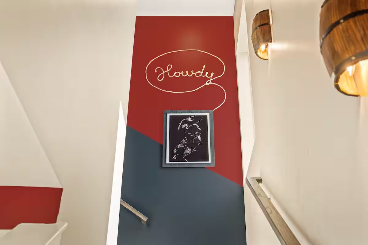 Interior staircase with red and navy geometric accent wall, cursive "Howdy" mural inside a rope-shaped speech bubble, framed black-and-white sketch and rustic barrel wall sconces