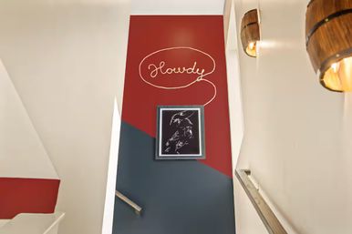 Interior staircase with red and navy geometric accent wall, cursive "Howdy" mural inside a rope-shaped speech bubble, framed black-and-white sketch and rustic barrel wall sconces