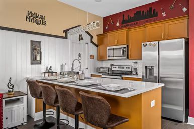 Nashville-themed modern kitchen with white quartz island and four brown bar stools, stainless steel fridge and oven, warm wood cabinets, and a red accent wall featuring a city skyline decal and decorative guitars.