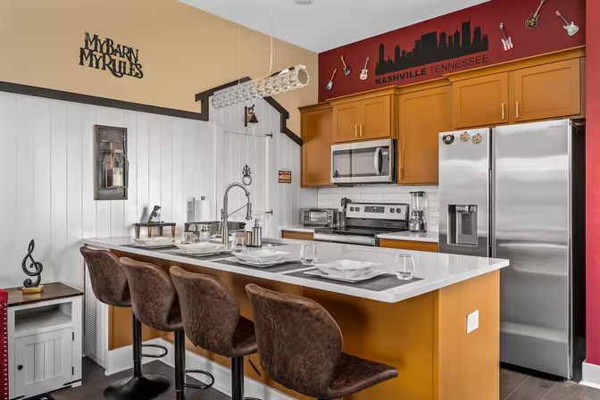 Nashville-themed modern kitchen with white quartz island and four brown bar stools, stainless steel fridge and oven, warm wood cabinets, and a red accent wall featuring a city skyline decal and decorative guitars.