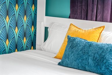 Vibrant bedroom close-up with white linens, teal Art Deco wallpaper, mustard yellow and teal velvet throw pillows and a white headboard