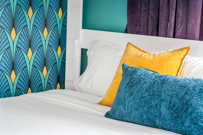 Vibrant bedroom close-up with white linens, teal Art Deco wallpaper, mustard yellow and teal velvet throw pillows and a white headboard