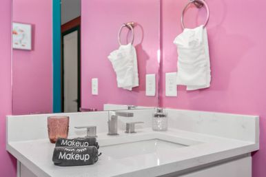 Bright pink boutique-style bathroom vanity with white quartz countertop, undermount sink and modern chrome faucet, mirror, towel ring with white hand towel, glass tumbler and stacked gray towels labeled "Makeup".