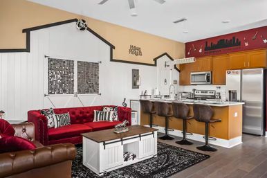 Open-concept Nashville, Tennessee living room and kitchen with red velvet tufted sofa, farmhouse white coffee table, barn-style wall accents, four brown leather bar stools at a breakfast bar, stainless steel appliances and skyline mural.