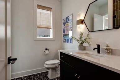 Bright modern powder room with black vanity and white countertop, octagonal mirror, black faucet, geometric black-and-white floor tile, white toilet, bamboo window shade, wall sconce, colorful wall art and vase of greenery.