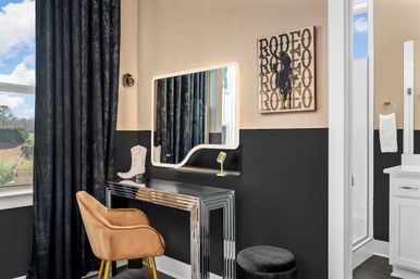 Stylish dressing nook adjacent to a bathroom with an illuminated mirror above a chrome console vanity, tan velvet chair, black ottoman, dark curtains, cowboy boot décor and rodeo wall art on two-tone black and beige walls.