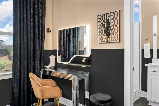 Stylish dressing nook adjacent to a bathroom with an illuminated mirror above a chrome console vanity, tan velvet chair, black ottoman, dark curtains, cowboy boot décor and rodeo wall art on two-tone black and beige walls.