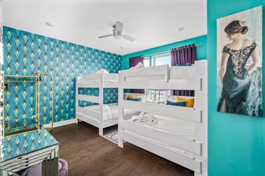 Bright kids' bedroom with two white twin bunk beds, teal art-deco wallpaper with gold accents, dark hardwood floor, purple curtains, colorful pillows and framed dress portrait.