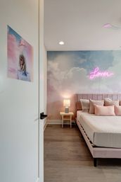 Dreamy pastel bedroom interior with cloud wall mural and pink neon "Lover" sign above a tufted pink upholstered bed, matching pillows, bedside table with lamp, hardwood floor, and a portrait poster on the open door.