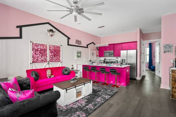 Bright open-concept living room and kitchen in a pink theme: hot-pink cabinets and island with stainless appliances, matching fuchsia sofa, black tufted loveseat and barstools, white shiplap accent wall with barn doors, large ceiling fan, and dark wood flooring.