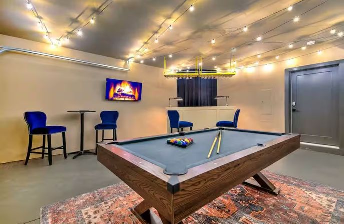 Cozy modern game room with a wooden pool table on a patterned rug, blue bar chairs, wall-mounted TV showing a fireplace, and string lights overhead.