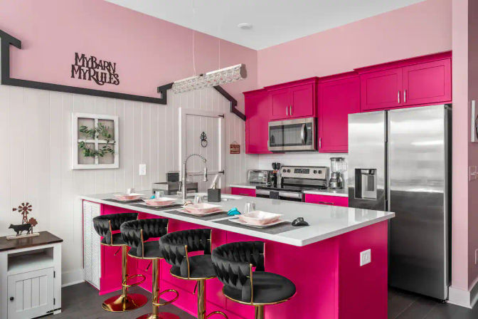 Vibrant hot-pink kitchen featuring a white countertop island, four black tufted bar stools with gold bases, stainless-steel appliances, pink cabinets and farmhouse-style accents.