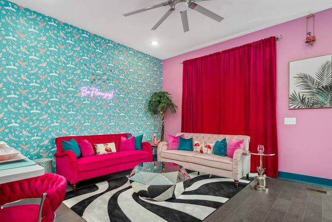 Vibrant pink-and-teal living room with flamingo-pattern wallpaper, neon wall sign, hot-pink and blush velvet sofas, mirrored geometric coffee table, black-and-white zebra rug, palm plant and bold red curtains.