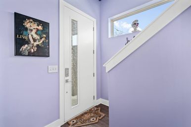 Bright lavender residential entryway with a modern white front door featuring a vertical glass insert, patterned doormat on dark hardwood floor, vintage floral portrait on the wall, and a playful decorative bust peeking through the stair window.